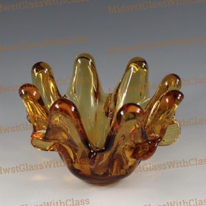 May include: Amber-colored glass bowl with a unique, organic shape. The bowl has a flared, petal-like design with multiple points extending outwards. The glass appears to be smooth and glossy, with variations in color from light to dark amber.