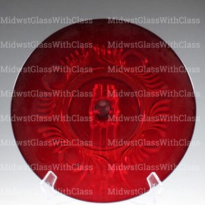 May include: A vibrant red glass plate with an ornate, embossed floral design. The plate is round and features a central raised area with a small, round detail. The words "MidwestGlassWithClass" are repeated around the edge.
