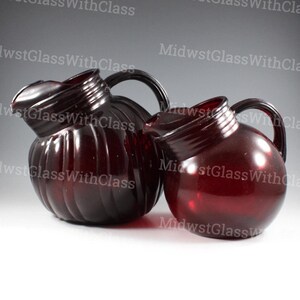 May include: Two ruby red glass pitchers with handles. The larger pitcher has a ribbed design, while the smaller one is smooth. Both have a spout for pouring. The pitchers are likely for serving beverages.