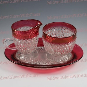 May include: A vintage glass sugar and creamer set with a ruby red trim and diamond pattern. The set includes a creamer with a handle, a sugar bowl, and a serving tray. The glass is clear with a red rim.