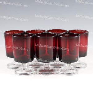 May include: A set of eight ruby red glass cordial glasses. Each glass features a cylindrical bowl and a clear, stemmed base. The glasses are arranged in a semi-circle, showcasing their uniform design and color. The background is a soft, neutral tone.
