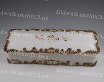 May include: A rectangular white trinket box with a gold-colored ornate trim and floral accents. The box features a hinged lid and is likely made of glass or porcelain. The floral design includes pink, yellow, and green flowers.