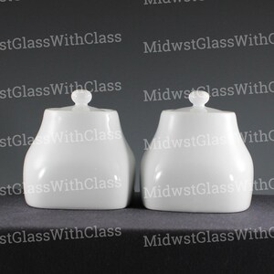May include: Two matching white ceramic sugar bowls with lids. Each bowl has a rounded body and a small knob on top of the lid. The bowls are sitting on a dark surface.