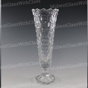 Puede incluir: Un jarrón de vidrio transparente con un patrón geométrico texturizado. El jarrón tiene una parte superior acampanada con un borde festoneado y una base hexagonal. El jarrón mide aproximadamente 25 cm de alto.