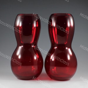 May include: Two ruby red glass vases with a unique hourglass shape. The vases have a glossy finish and are displayed against a neutral background. The overall design is simple yet elegant, suitable for home decor.