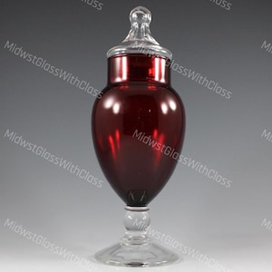 May include: A ruby red glass apothecary jar with a clear glass lid and base. The jar has a bulbous body and a clear, rounded stopper. The base is a clear, circular foot. The jar is likely used for storing items.
