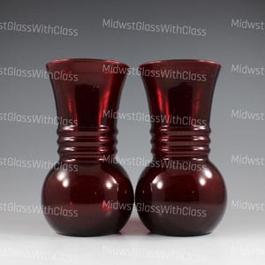 May include: Two ruby red glass vases. Each vase has a bulbous base, a ridged midsection, and a flared top. The vases are identical in shape and color, with a glossy finish. The background is a gradient of gray and white.
