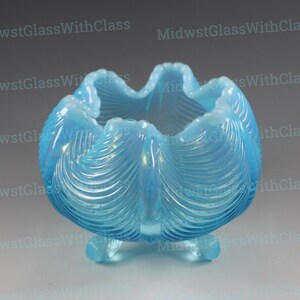 May include: A light blue, opalescent glass bowl with a ruffled rim and three small feet. The bowl has a textured, shell-like pattern on the exterior. The text "MidwestGlassWithClass" is visible in the background.