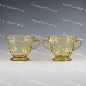 May include: A pair of vintage yellow glass serving dishes. One is a creamer with a spout and handle, the other is a sugar bowl with two handles. Both have a floral pattern and sit on a pedestal base.