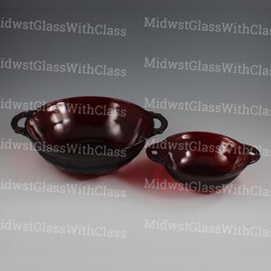 May include: Two ruby red glass bowls with handles. The larger bowl has a textured design around the base. Both bowls are on a light background. The bowls are likely for serving.