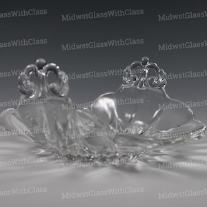 May include: Clear glass decorative bowl with a scalloped edge and two ornate, crown-like handles. The bowl has a shallow, open design and sits on a reflective surface. The glass is transparent, allowing light to pass through.