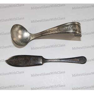 May include: Two antique silver serving utensils: a spoon with "Cream Top" engraved on the handle and a butter knife. The spoon has a rounded bowl and a curved handle. The butter knife has a flat, wide blade and a slender handle.