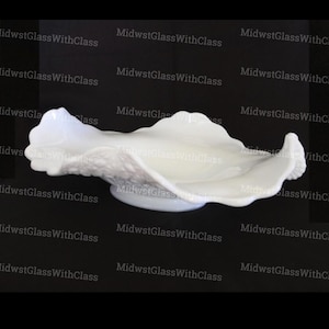 May include: A white, opalescent glass serving dish with a ruffled, shell-like design. The dish has a wide, shallow bowl and a raised, circular base. The edges are scalloped, and the surface has a textured pattern. The background is black.