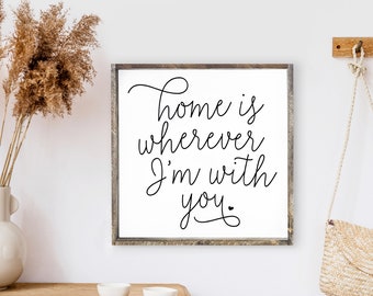 May include: A white framed sign with black calligraphy that reads "Home is wherever I'm with you."  The sign is hanging on a white wall with a wooden shelf below it.