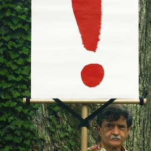 May include: A white banner with a large red exclamation point, held by a person. The banner is mounted on wooden dowels and supported by black straps. The person is wearing a patterned shirt.