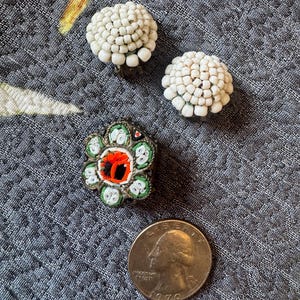 Peut inclure: Deux boucles d'oreilles clips en forme de dôme recouvertes de perles blanches et une broche mosaïque florale composée de carreaux de verre verts, blancs et orange. Une pièce de 25 cents américaine est placée à côté pour donner l'échelle.