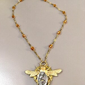 Puede incluir: Un collar dorado con un colgante de abeja detallado. El colgante de abeja tiene un centro transparente que revela intrincados detalles de relojería. La cadena del collar está adornada con cuentas de color ámbar, creando un accesorio único y llamativo.