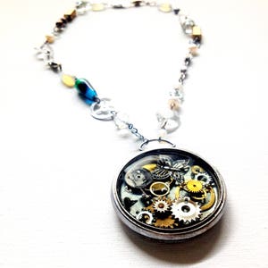 May include: A silver necklace with a round pendant featuring a clear resin filled with gears and a butterfly. The chain is made of silver and has clear crystal beads and gold accents.