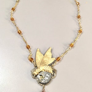 Puede incluir: Un collar de oro con un colgante de estilo steampunk que presenta una mariposa con engranajes y un cristal de ámbar en forma de lágrima. El collar está hecho con una cadena de oro y cuentas de ámbar.