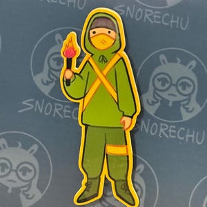 May include: A sticker featuring a cartoon figure in a green hooded outfit with yellow accents, holding a torch with a flame. The figure wears a yellow mask. The background has the word "SNORECHU" repeated.