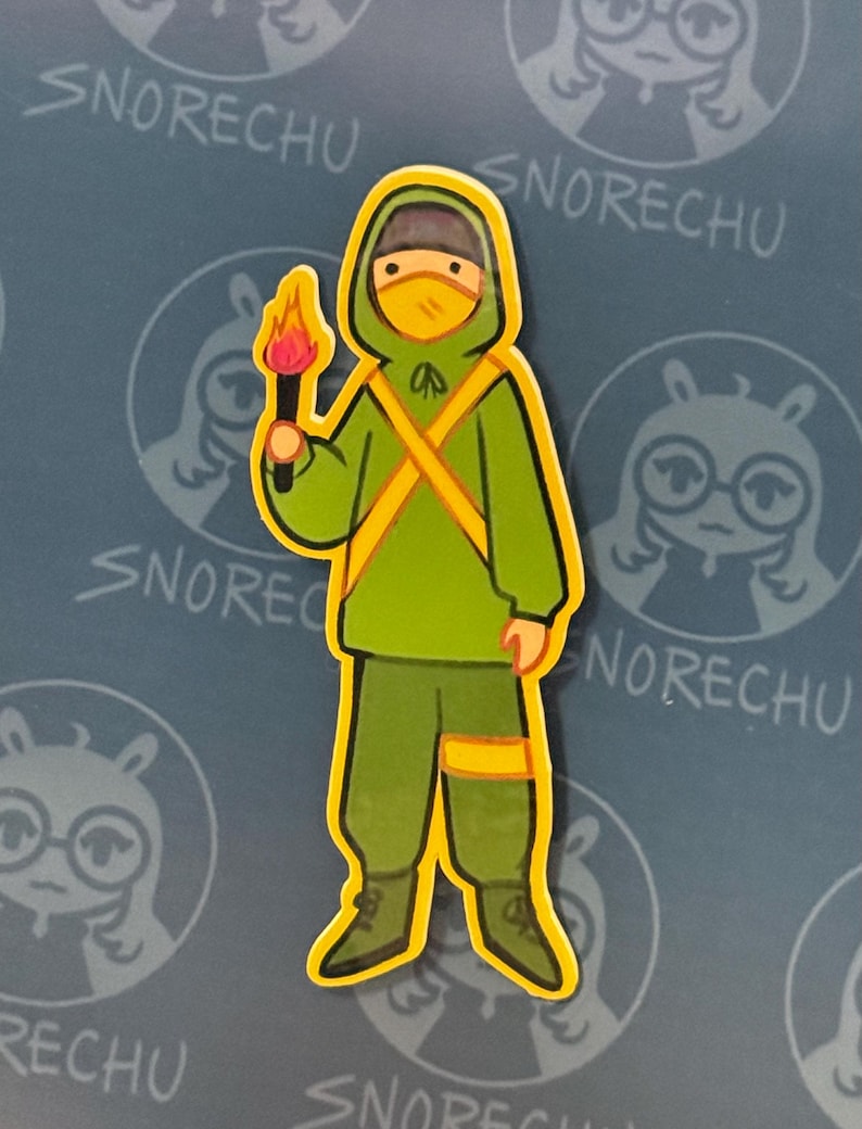 May include: A sticker featuring a cartoon figure in a green hooded outfit with yellow accents, holding a torch with a flame. The figure wears a yellow mask. The background has the word "SNORECHU" repeated.
