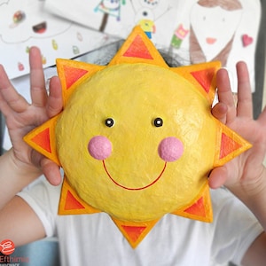 Op de afbeelding: Een kind houdt een gele papier-maché zon vast met een glimlachend gezicht, rode stralen en roze wangen. De zon is een handgemaakt ambacht.