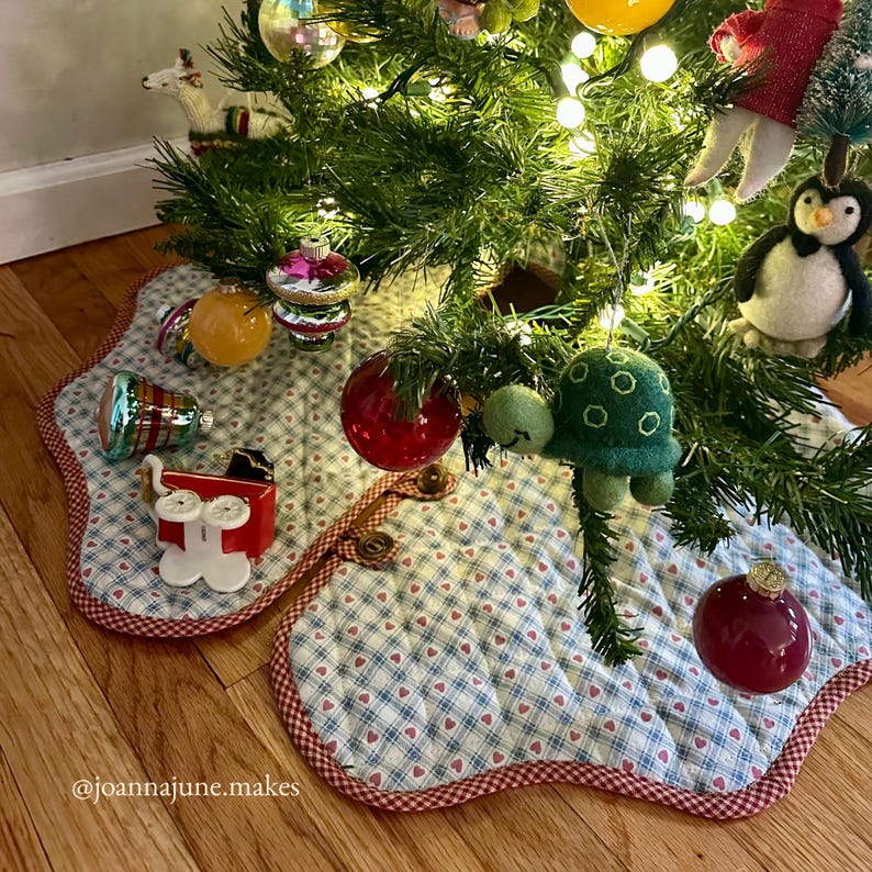 May include: A quilted Christmas tree skirt with a blue and white plaid pattern and red heart accents. The skirt is trimmed with a red border and features a button closure. Ornaments and a felt turtle are on the tree.