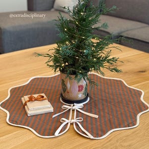 May include: A small potted evergreen tree with string lights sits on a decorative, scalloped-edge tree skirt. The tree skirt is brown and gray striped with a white border and bow ties. Two wrapped gifts with a brown ribbon are on the skirt.
