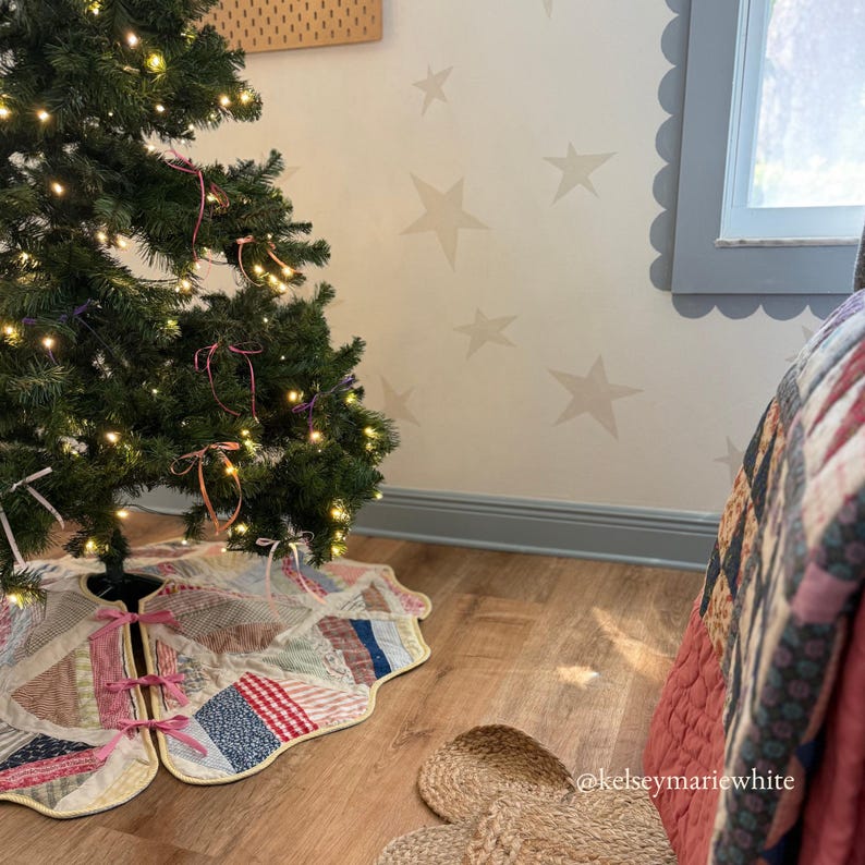 May include: A decorated Christmas tree with a patchwork tree skirt. The tree skirt is made of various fabric patches in red, blue, and cream colors, with pink bows. The tree is lit with warm white lights and has pink and gold ribbon accents. The background features a star-patterned wall.