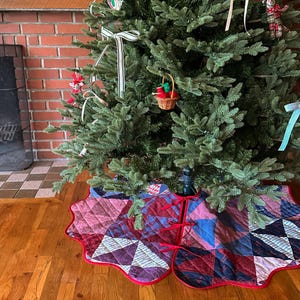 May include: A decorated Christmas tree with a quilted tree skirt. The tree skirt is made of patchwork fabric in shades of red, blue, and purple, with a scalloped edge. The tree is adorned with ornaments and ribbons.