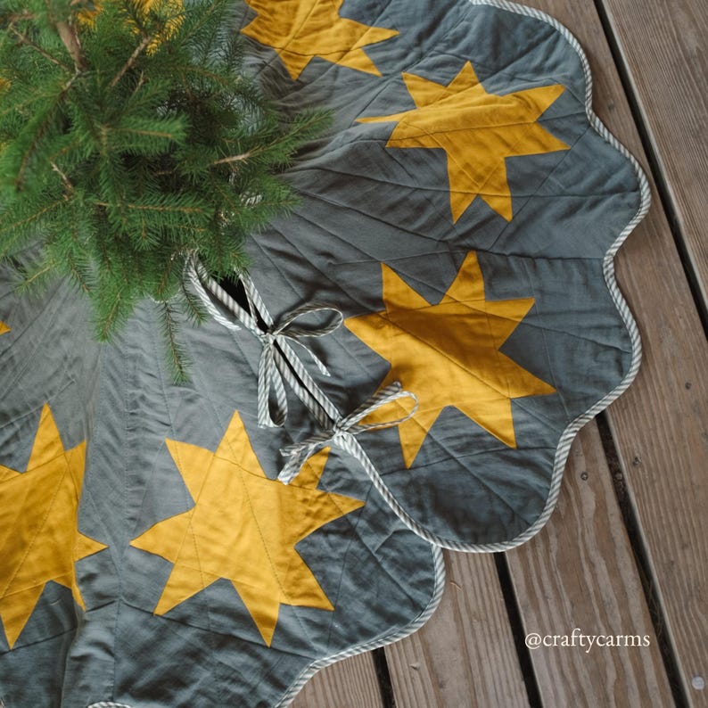 May include: A gray and gold star-patterned Christmas tree skirt with a scalloped edge and rope trim. The skirt has a quilted design and is tied with gray and white striped ribbons. A small evergreen tree is partially visible.
