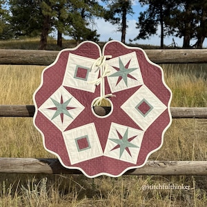 May include: A quilted Christmas tree skirt with a scalloped edge. The skirt features a burgundy background with cream-colored squares and teal star designs. A cream-colored tie is at the top. The skirt is approximately 48 inches in diameter.