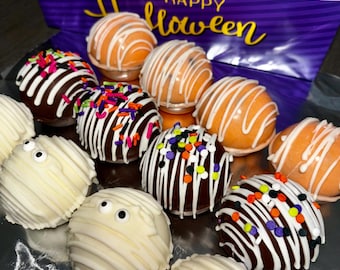 May include: A box of Halloween-themed chocolate truffles. Some are decorated with white icing stripes, orange icing, and colorful sprinkles. Others are designed to look like mummies with white icing and candy eyes. The box has a purple and white design with the words "Happy Halloween".