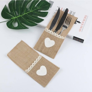 May include: Two burlap cutlery holders with white lace trim and a white heart applique. One holder contains a knife, fork, and spoon. The other holder is empty. The holders are on a white surface.
