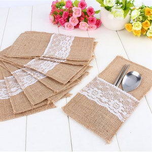 May include: A stack of burlap place card holders with white lace trim. The holders are designed to hold silverware or other small items.