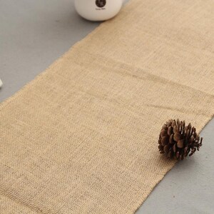May include: A natural brown burlap table runner with a pine cone on a grey surface.