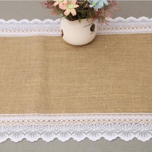 Puede incluir: Un camino de mesa de yute marrón con un borde de encaje blanco. El camino de mesa está decorado con un pequeño ramo de flores blancas y rosas.