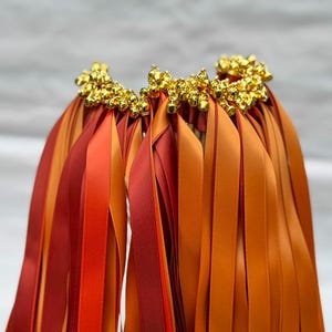 May include: Close-up of a decorative item with multiple strands of orange and red ribbons. The ribbons are topped with clusters of small, gold-colored beads. The ribbons are arranged in a fan-like shape.