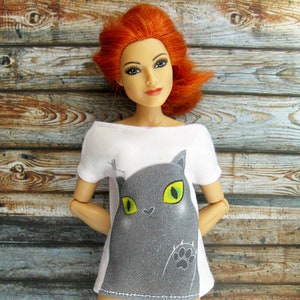 Pode incluir: Uma camiseta branca com um gr&aacute;fico de gato cinza com olhos verdes. O gato est&aacute; olhando para o espectador e tem uma pegada na pata.