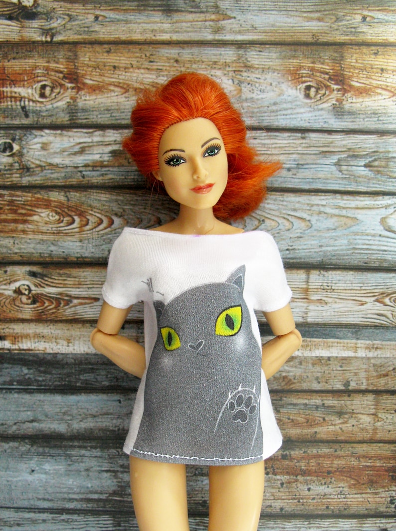 Pode incluir: Uma camiseta branca com um gr&aacute;fico de gato cinza com olhos verdes. O gato est&aacute; olhando para o espectador e tem uma pegada na pata.