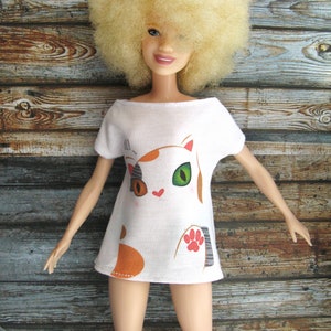 Pode incluir: Uma camiseta branca com um desenho de gato de desenho animado. O gato tem p&ecirc;lo laranja, branco e preto e olhos verdes. A camiseta est&aacute; sendo usada por uma boneca com uma peruca afro loira.