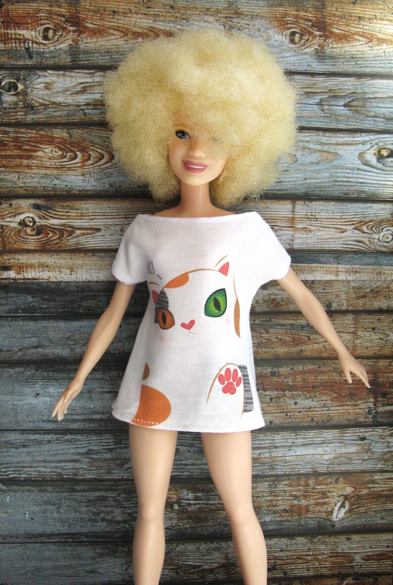 Pode incluir: Uma camiseta branca com um desenho de gato de desenho animado. O gato tem p&ecirc;lo laranja, branco e preto e olhos verdes. A camiseta est&aacute; sendo usada por uma boneca com uma peruca afro loira.