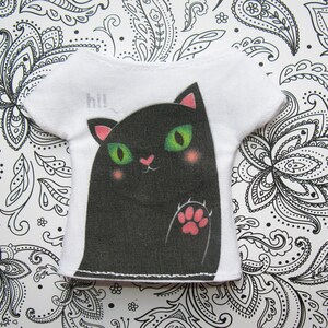 Pode incluir: Camiseta branca com um gr&aacute;fico de um gato preto e a palavra "hi!" impressa na frente.