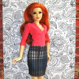 May include: A red and black plaid skirt with a pink long-sleeved shirt. The doll has long red hair and is wearing a pink shirt with a v-neck.