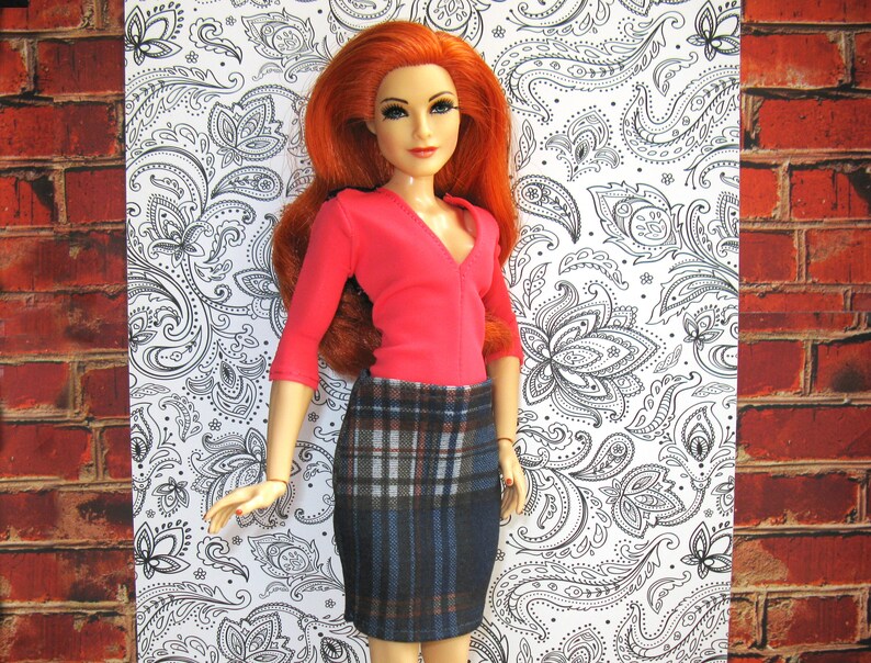 May include: A red and black plaid skirt with a pink long-sleeved shirt. The doll has long red hair and is wearing a pink shirt with a v-neck.