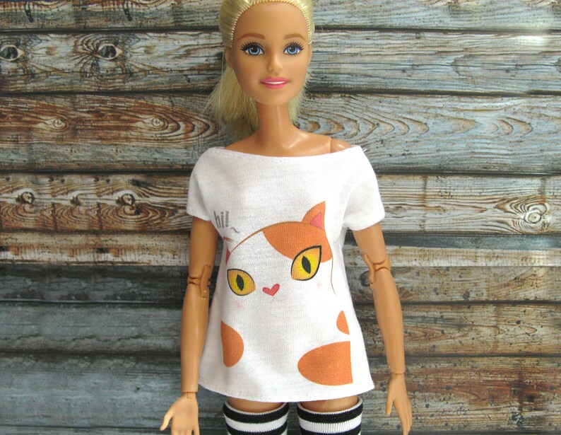 Pode incluir: Camiseta branca com um gr&aacute;fico de gato de desenho animado em laranja e branco. O gato tem um nariz de cora&ccedil;&atilde;o rosa e o texto "hill" est&aacute; acima do gato.