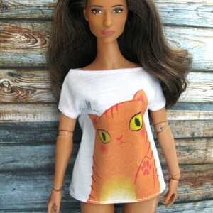 Pode incluir: Uma boneca vestindo uma camiseta branca com um gr&aacute;fico de gato laranja de desenho animado. O gato tem olhos amarelos e um nariz rosa.