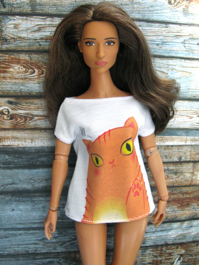 Pode incluir: Uma boneca vestindo uma camiseta branca com um gr&aacute;fico de gato laranja de desenho animado. O gato tem olhos amarelos e um nariz rosa.