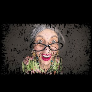 May include: A woman with grey hair and a surprised expression wears black-rimmed glasses and a green floral print blouse.
