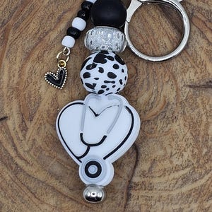 May include: A keychain featuring a stethoscope design, with a heart-shaped charm and various beads in black, white, and silver. The keychain has a silver clasp and key ring, perfect for medical professionals or students.